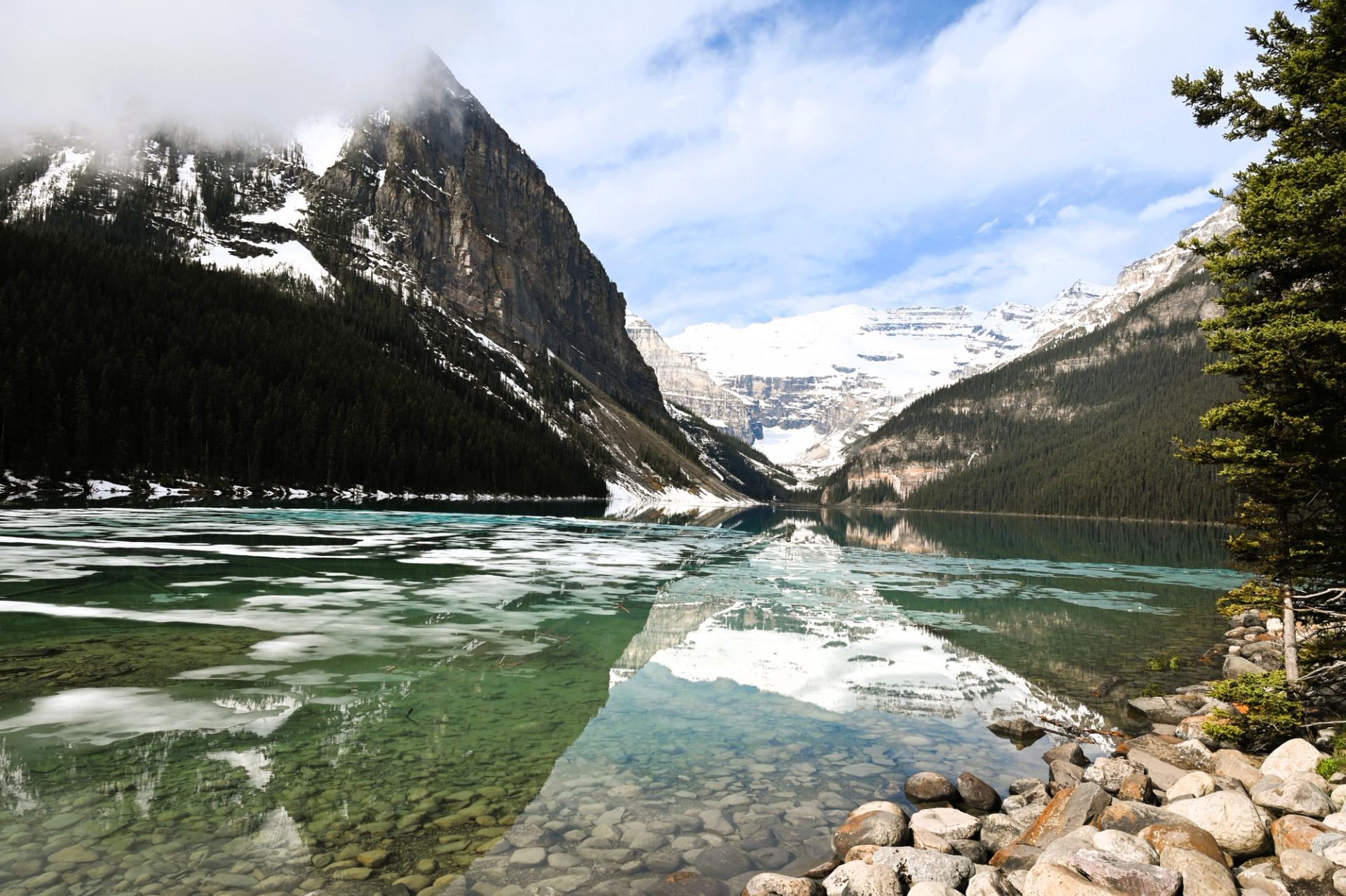 Rocky shoreline beside turquoise lake with floating ice, surrounded by snow-capped mountains.