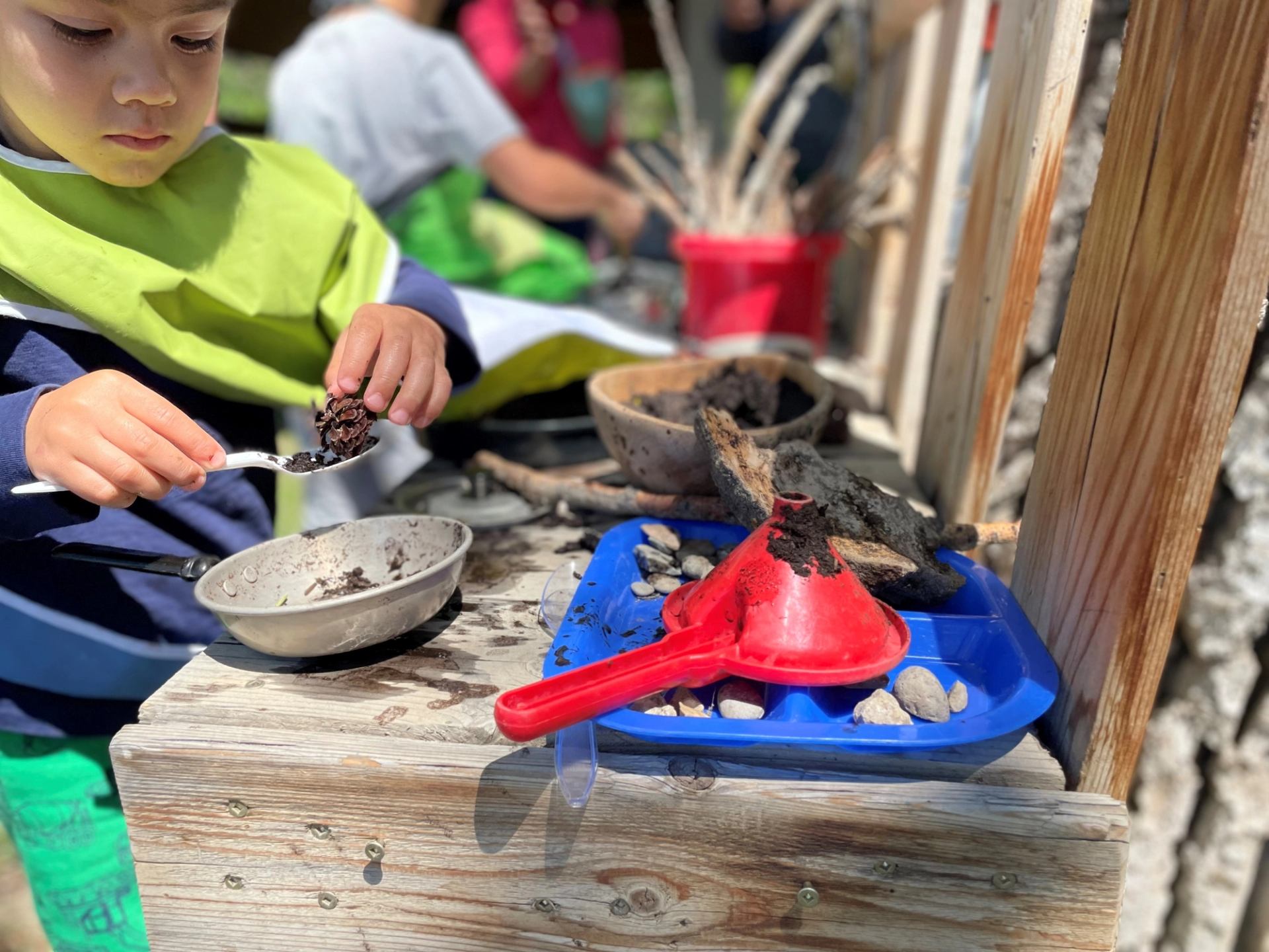 Child scoops soil into a pan beside toy funnel, tray, rocks and sticks.