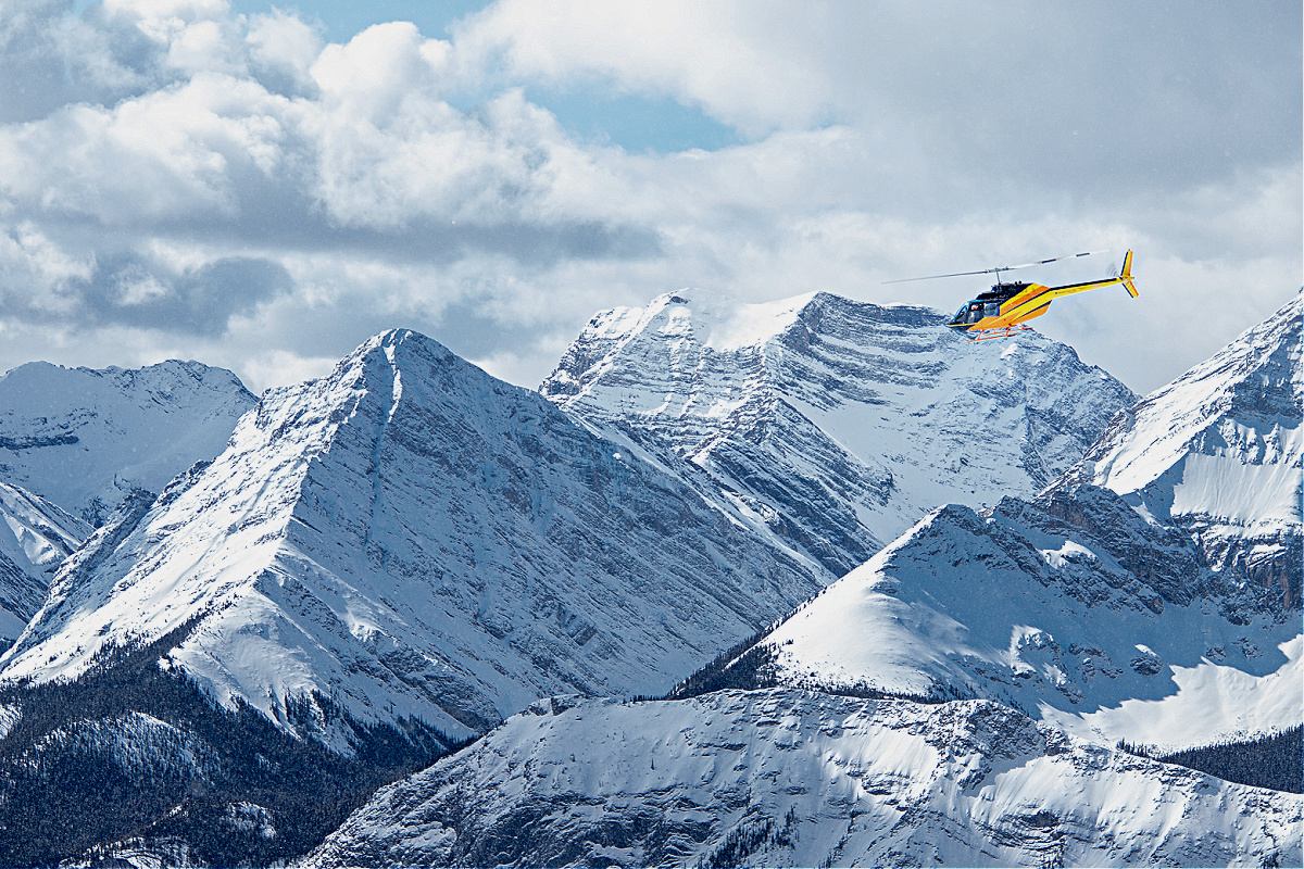 A helicopter flies past snow-covered mountain peaks under dramatic clouds.