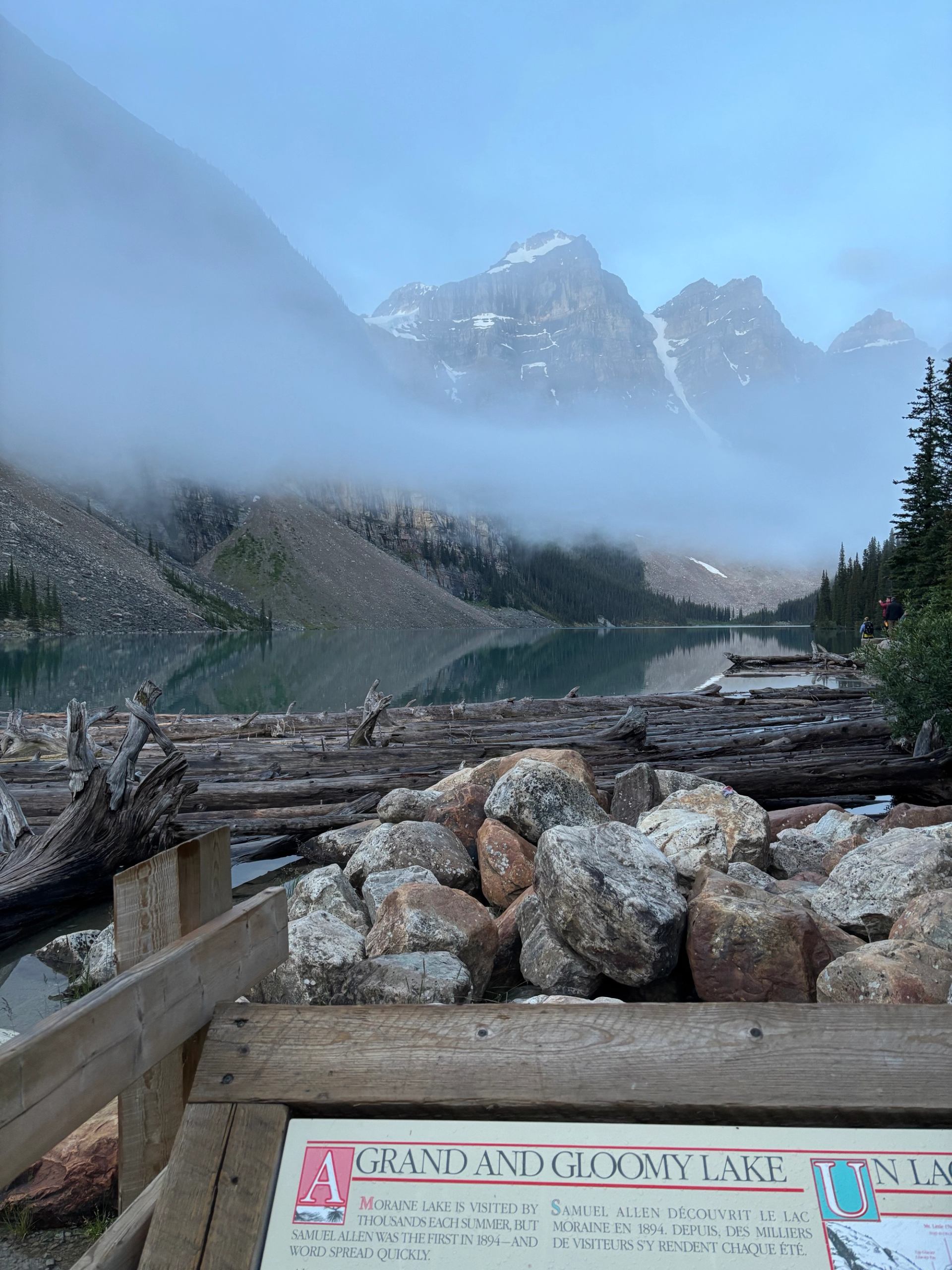 Foggy mountain lake with driftwood, rocky shoreline and wooden sign in foreground.