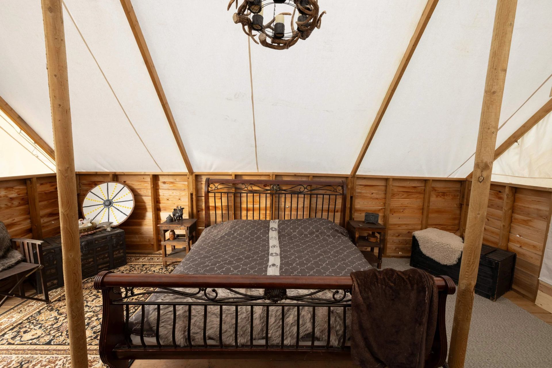 A luxurious glamping tent bedroom with a large bed, wooden walls, white canvas ceiling, and rustic decor.