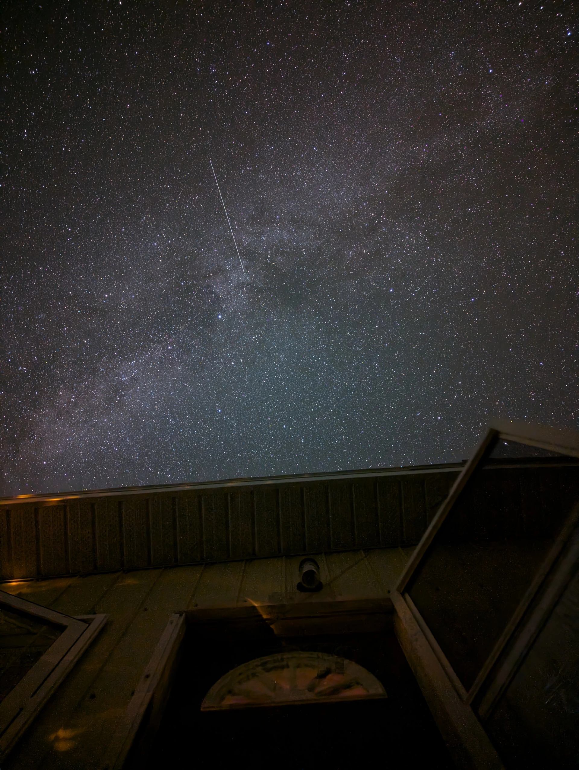 Star-filled night sky with a meteor above a building and open windows.