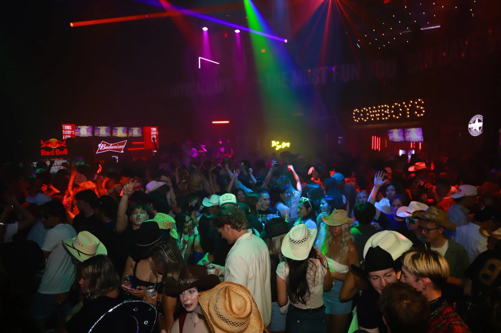 Crowded nightclub dance floor with people in cowboy hats under colourful stage lights.