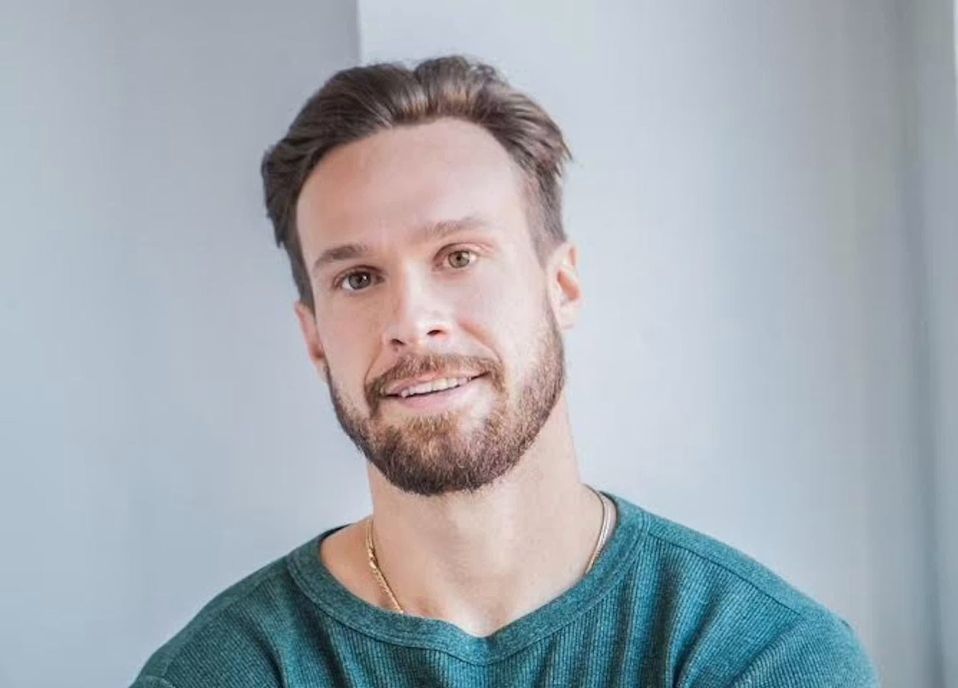 Bearded person in green sweater facing camera against plain light-coloured indoor wall.