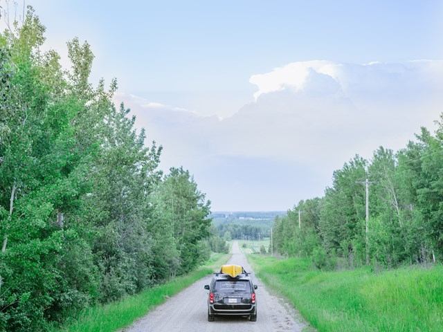 Campervan with a kayak on top driving through a forested dirt road under a clear sky.