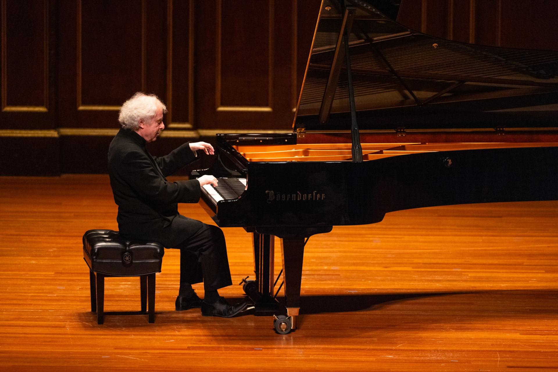 Pianist in black suit plays Bösendorfer grand piano on polished wooden concert stage.