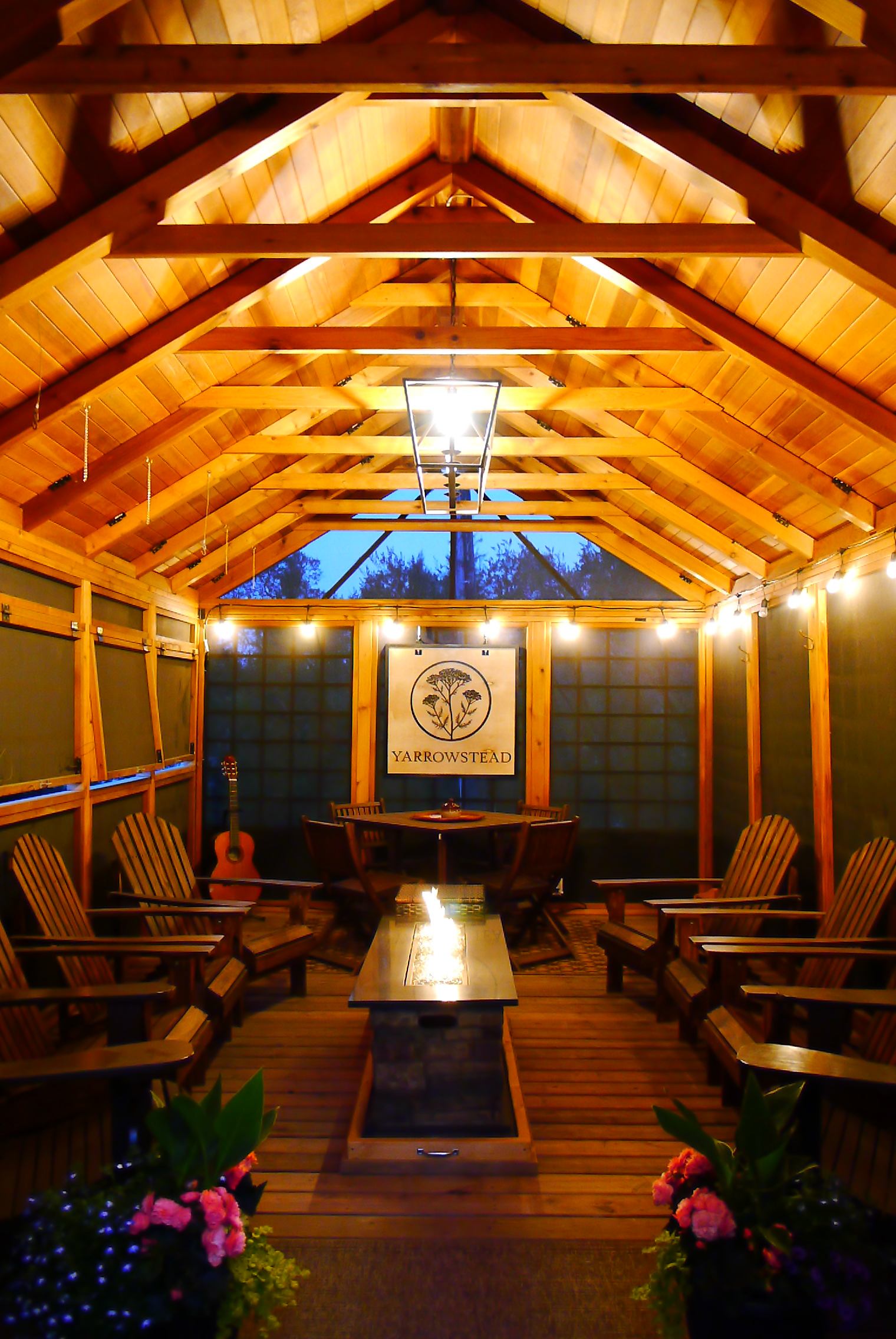 A cozy enclosed lounge features wooden beams, Adirondack chairs, and a long fire table in the center. Soft lighting and a “Yarrowstead” sign on the wall add warmth to the inviting relaxation space.