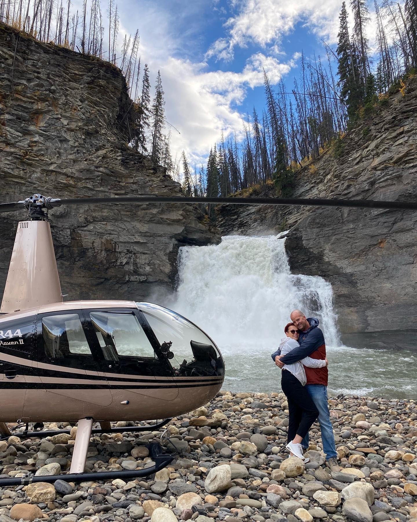 Helicopter near waterfall with couple embracing on rocky terrain.
