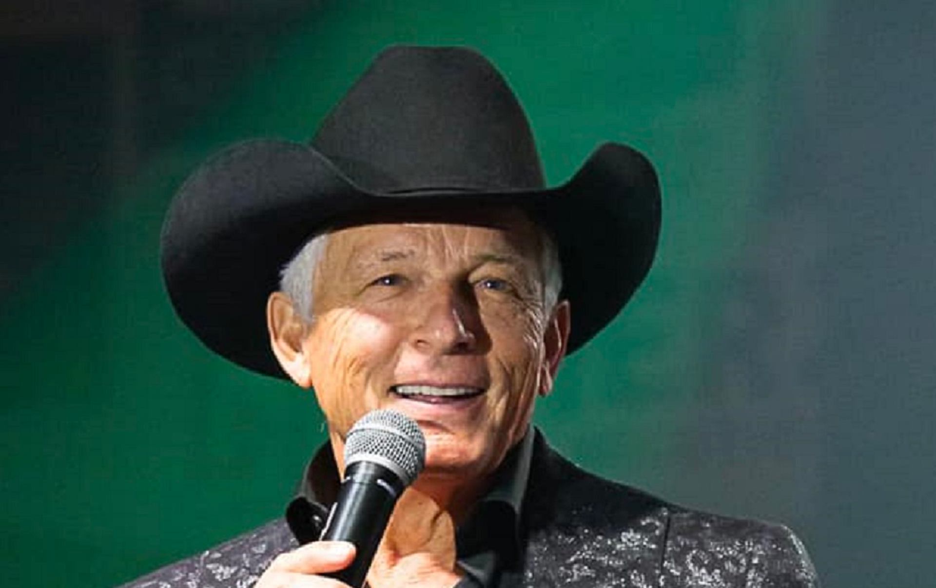Country singer in black cowboy hat holds microphone on stage with green background.