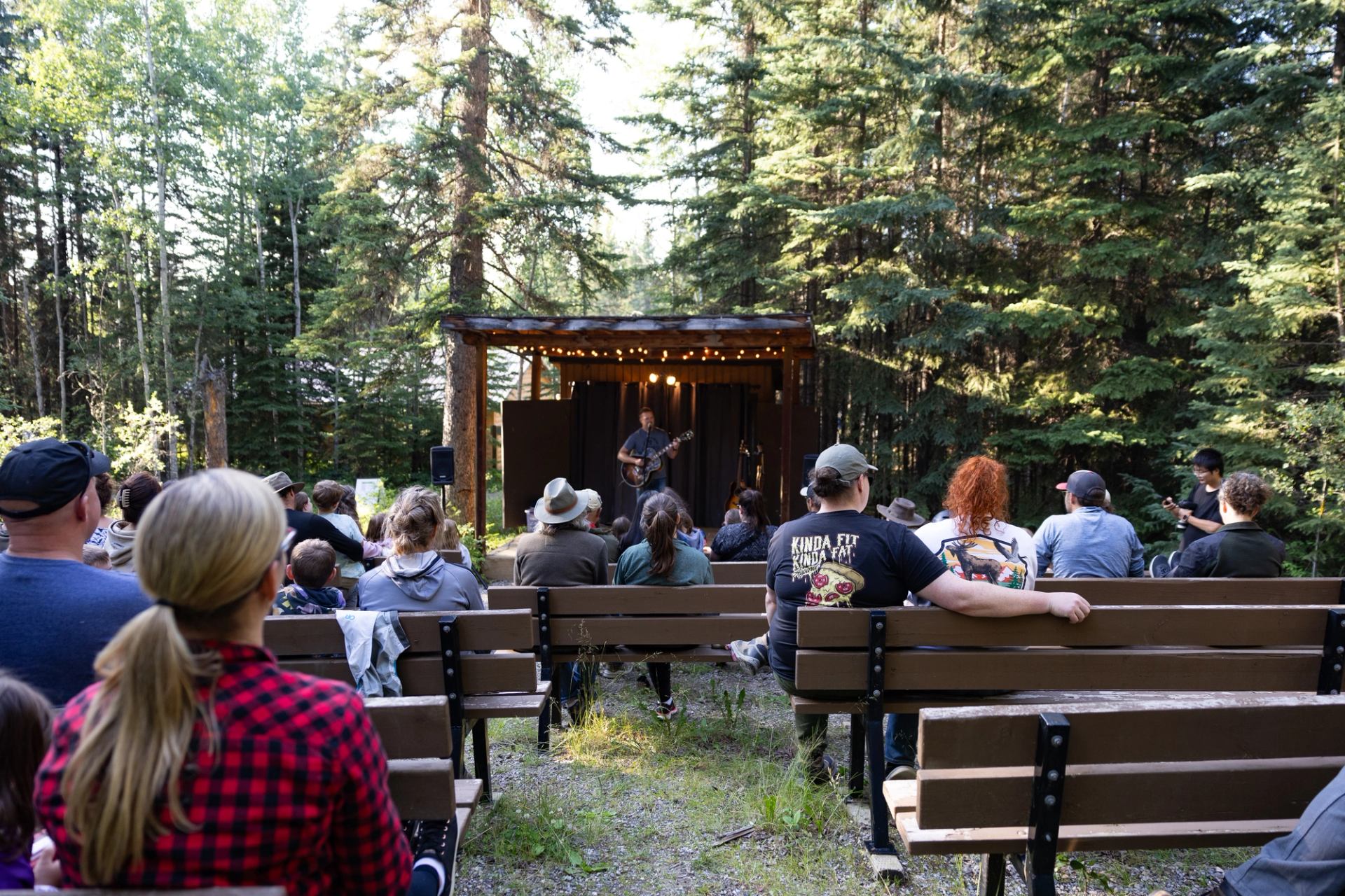 People seated on benches in forest watching outdoor stage with string lights.