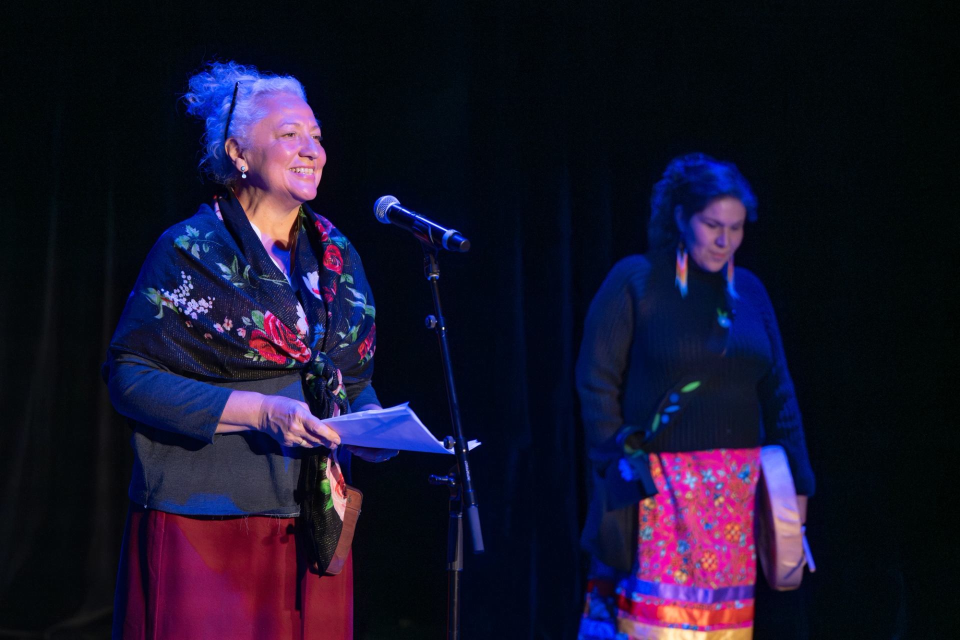 Two performers standing onstage, one speaking at a microphone and one holding a drum.