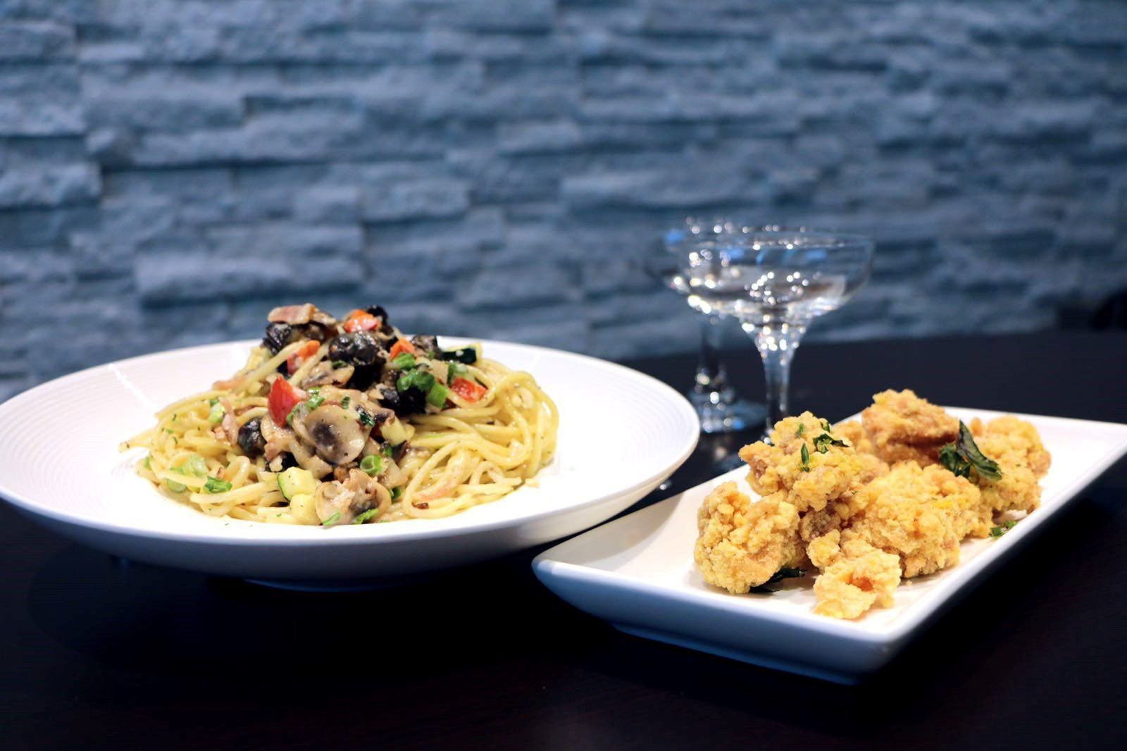 Plates of pasta with vegetables and fried chicken with a glass on a table.