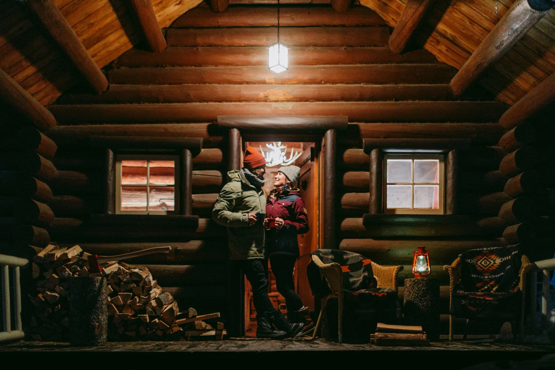 Two people stand on a lit porch of a log cabin at night.