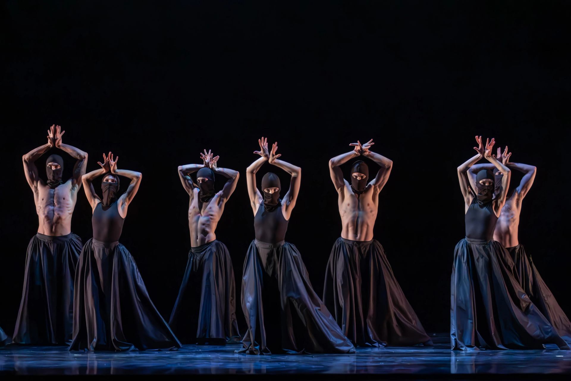 A group of ballet dancers in a line. They are dressed in black and have their arms over their heads.