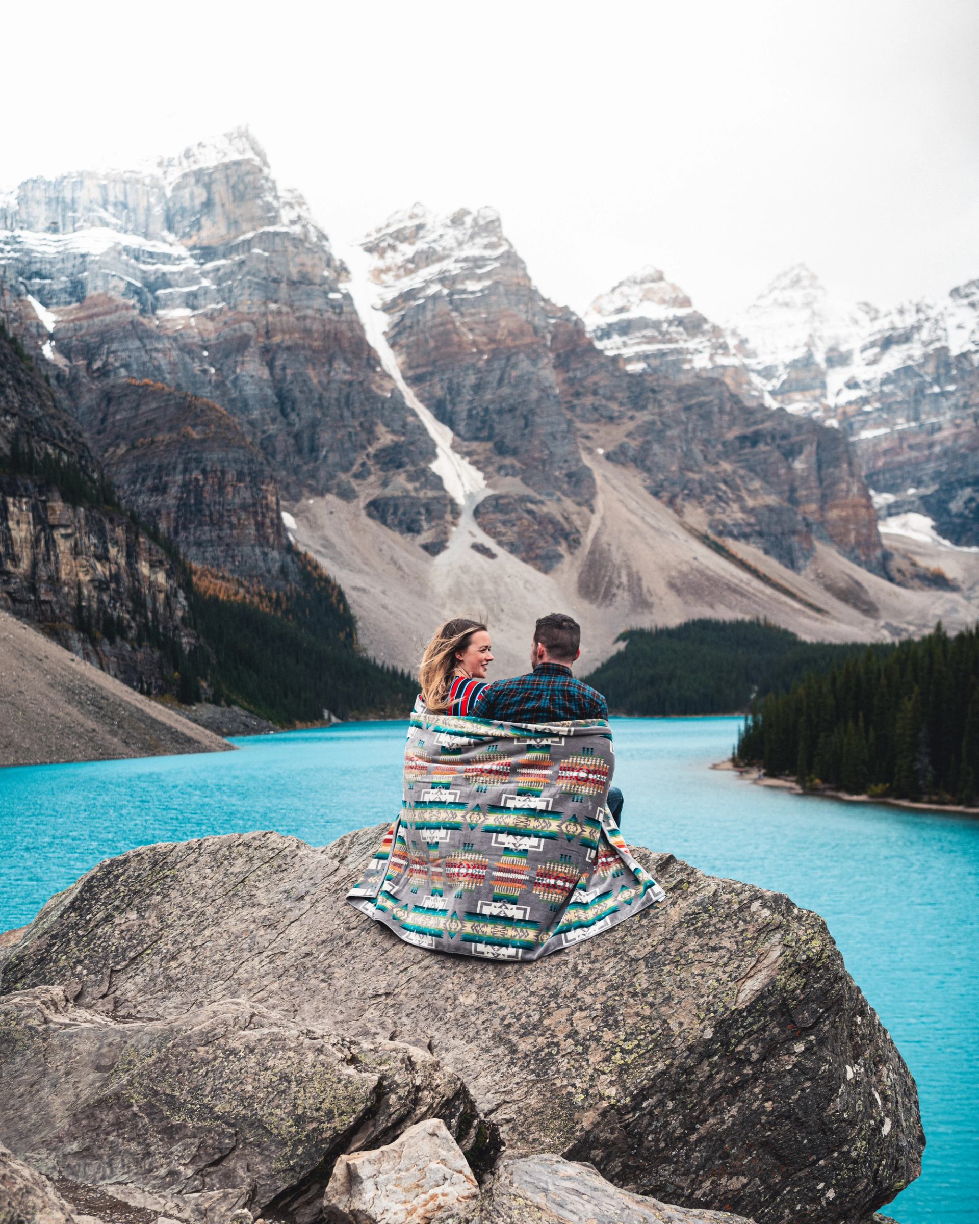 Two people wrapped in a blanket sit on lakeside rock below snowy mountains.