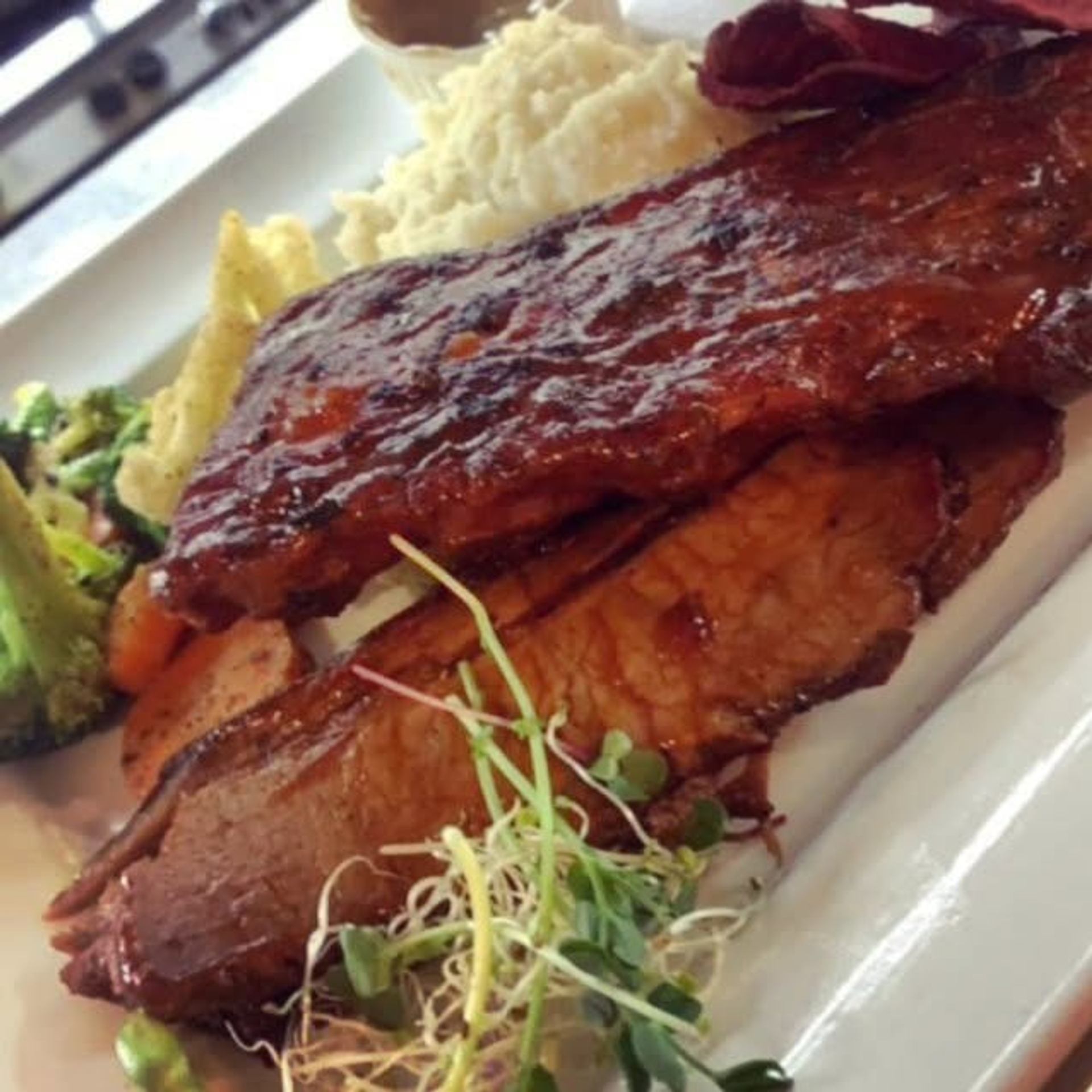 Barbecue ribs coated in glossy sauce are served with mashed potatoes, vegetables, and fresh sprouts.