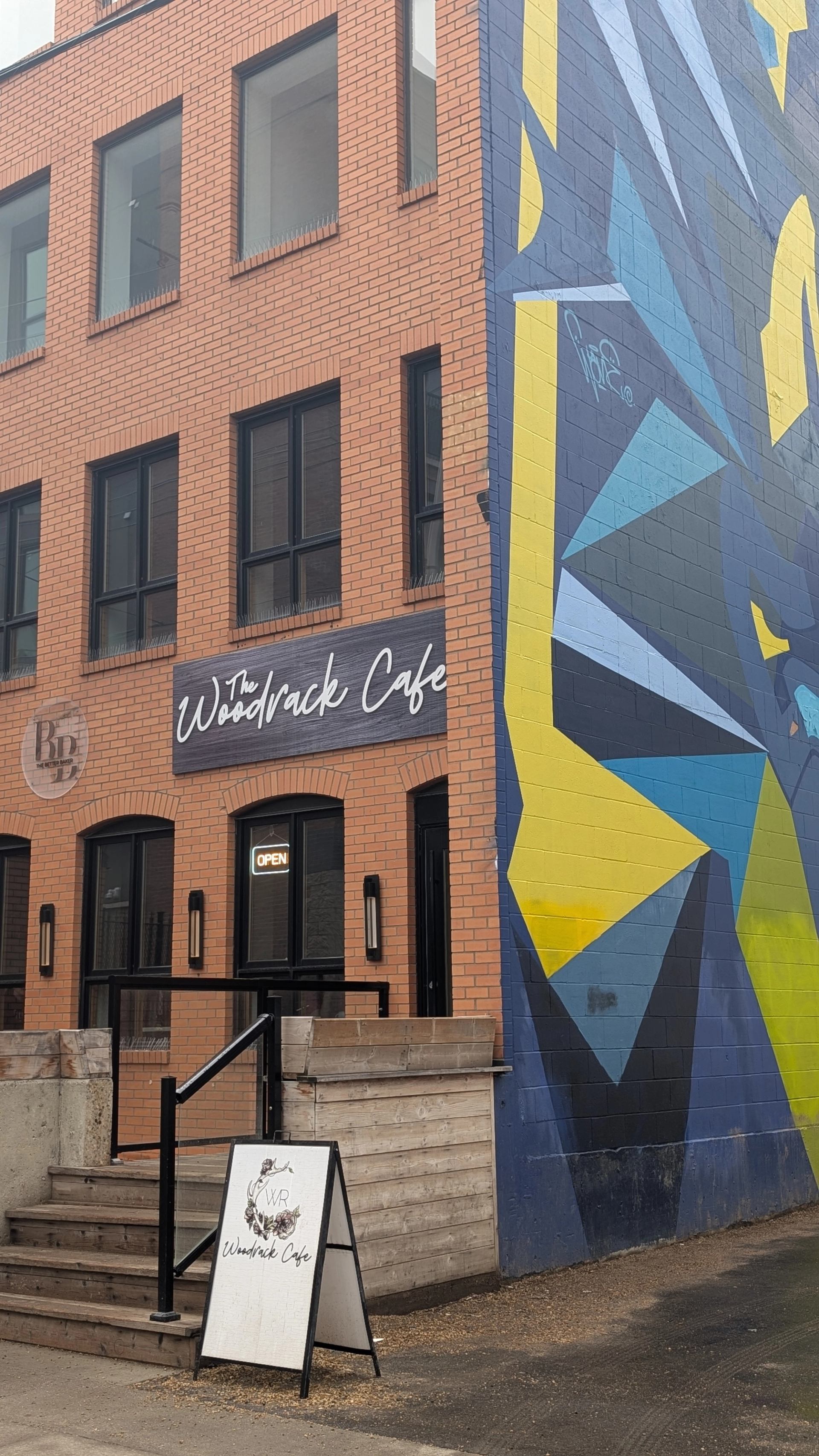 Brick building with The Woodrack Cafe sign beside large geometric mural and sandwich board.