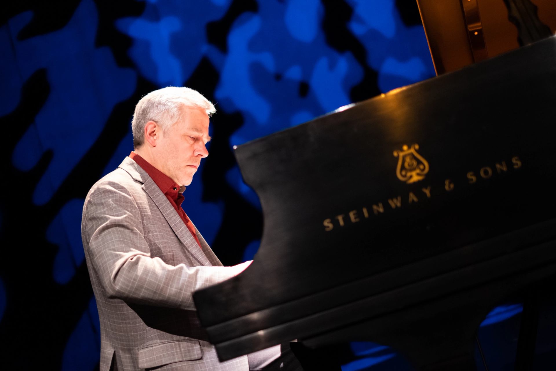 Pianist in grey blazer plays Steinway & Sons grand piano against blue abstract backdrop.