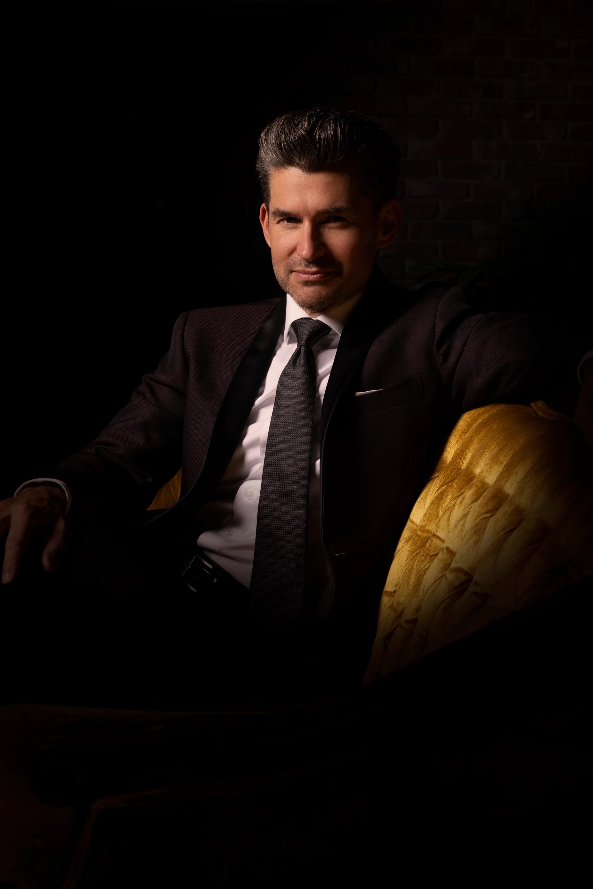 Man in dark suit sits on gold velvet chair in low studio lighting.