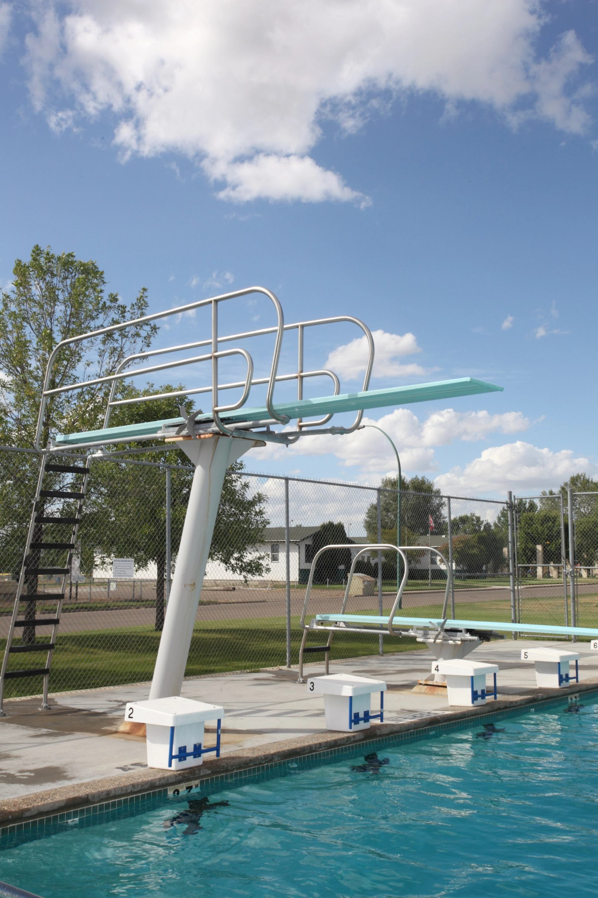 Swimming pool diving boards with chain-link fencing and blue sky with clouds.