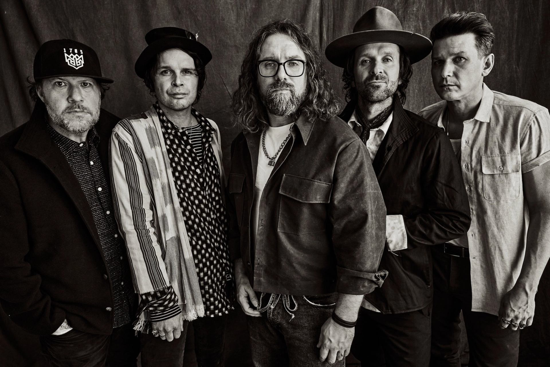 Five men in varied hats and casual jackets stand together for a studio portrait.