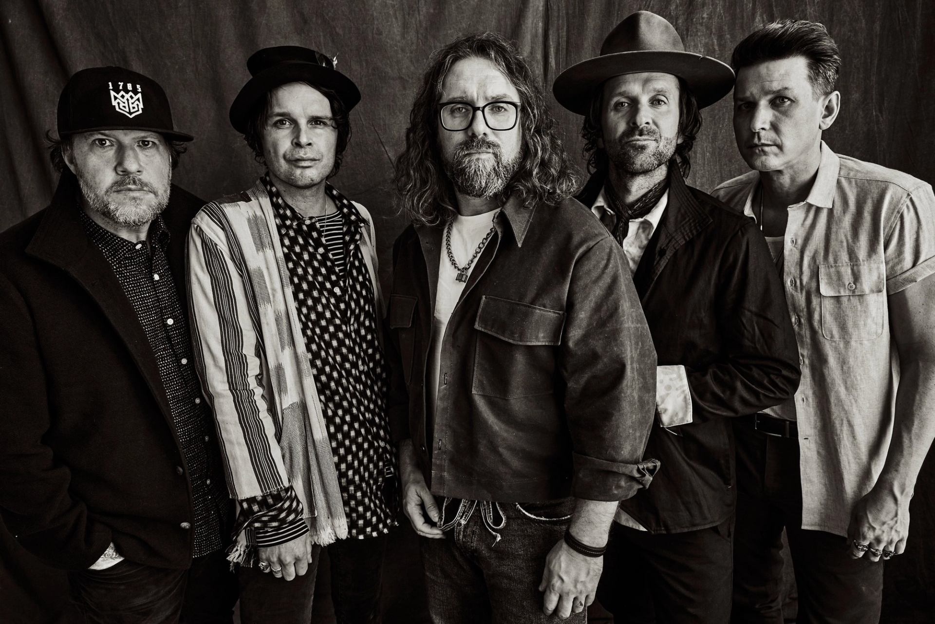 Five men in varied hats and casual jackets stand together for a studio portrait.