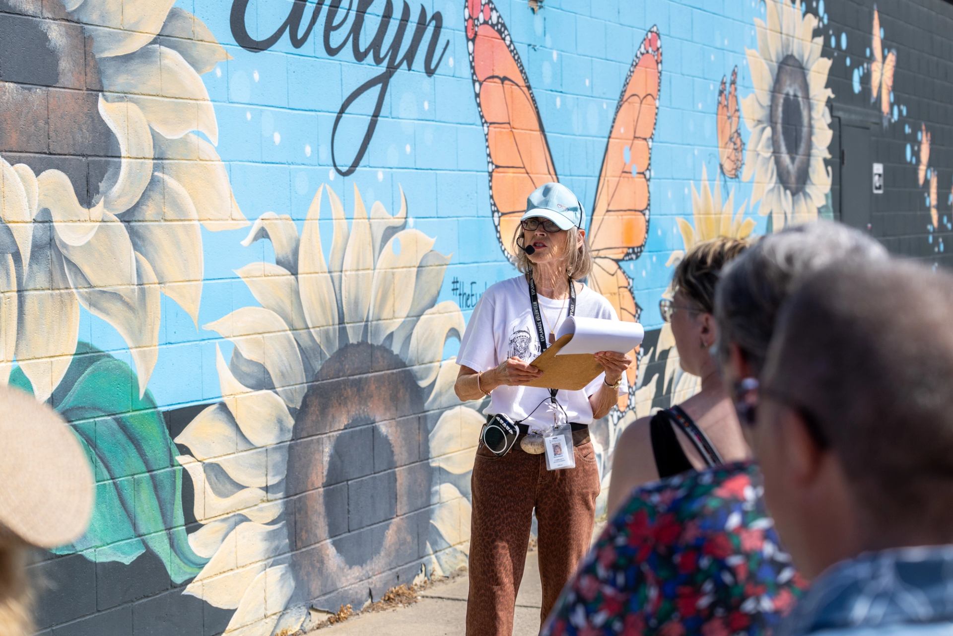 Tour guide with microphone speaks to group beside sunflower and butterfly mural.
