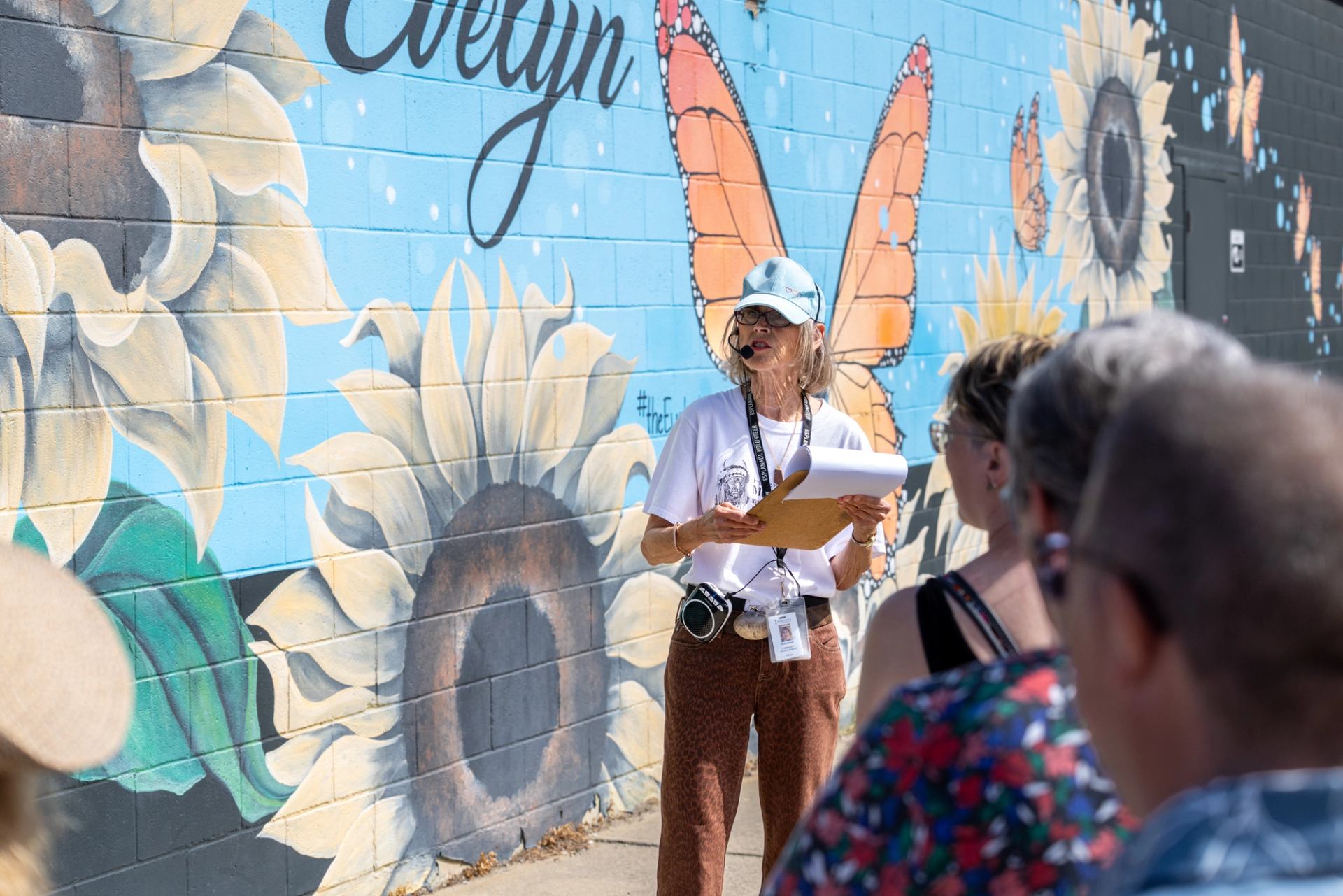 Tour guide with microphone speaks to group beside sunflower and butterfly mural.