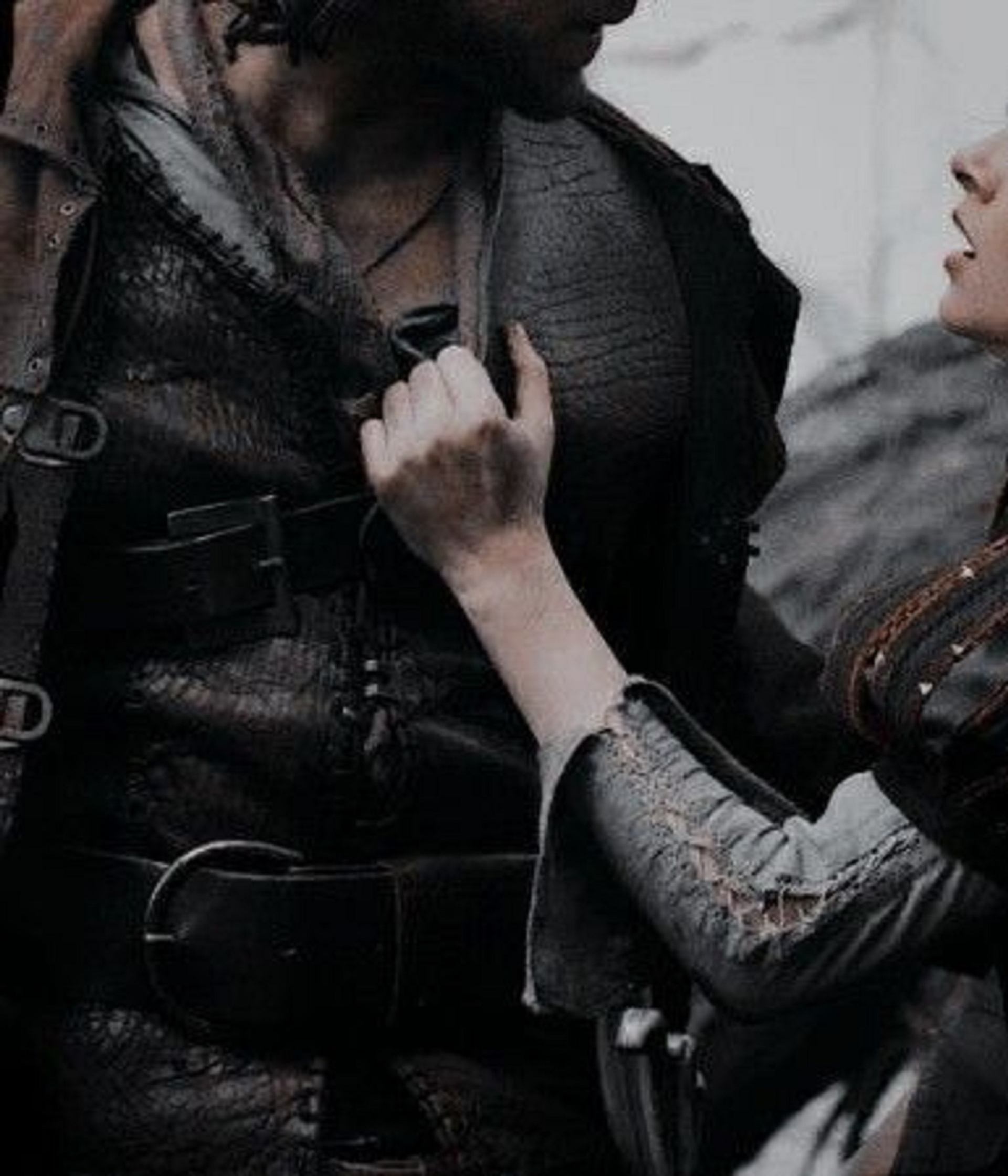 Woman's hand on a man's chest, both in dark, rugged clothing. Her mouth is slightly open as she looks up at him.