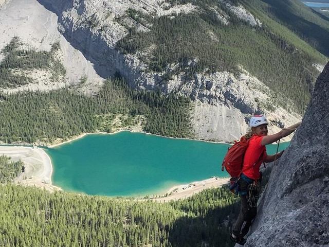 A climber in a red shirt ascends a rocky cliff, turquoise lake and green forest below, full of adventure and grit.