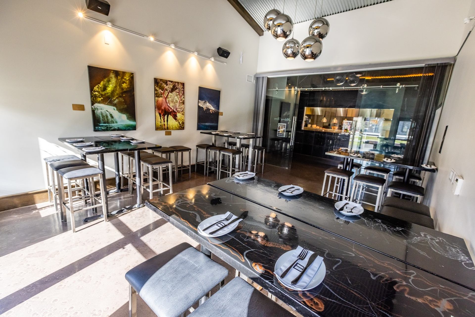A modern restaurant dining room with high-top tables, a long dark communal table, nature artwork, and a glass-enclosed kitchen.