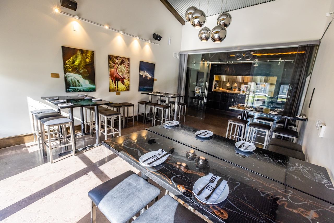 A modern restaurant dining room with high-top tables, a long dark communal table, nature artwork, and a glass-enclosed kitchen.