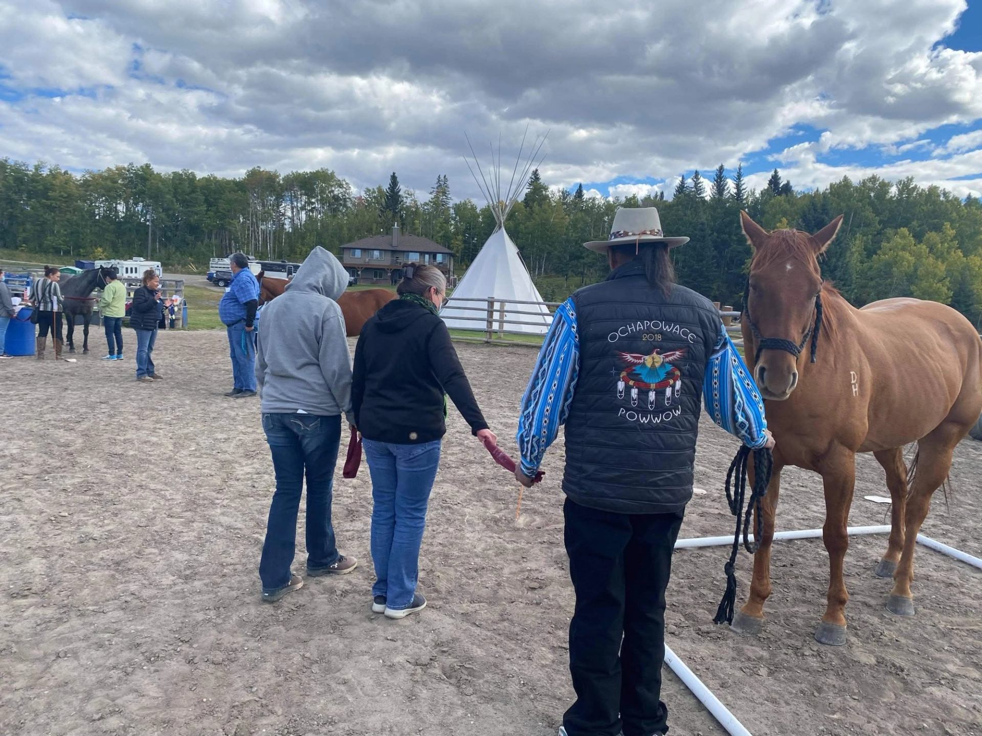 People stand with horses in an outdoor corral near a teepee and trees.