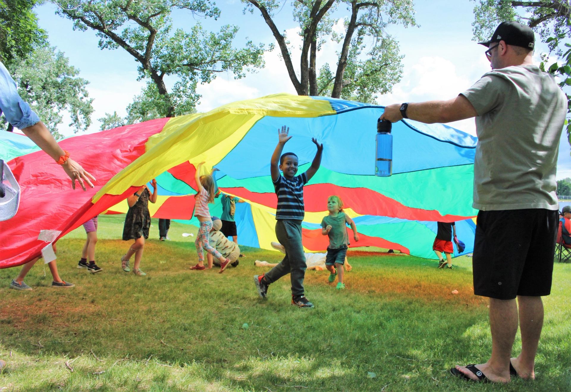 Children run under a large colourful parachute held by adults in a grassy park.