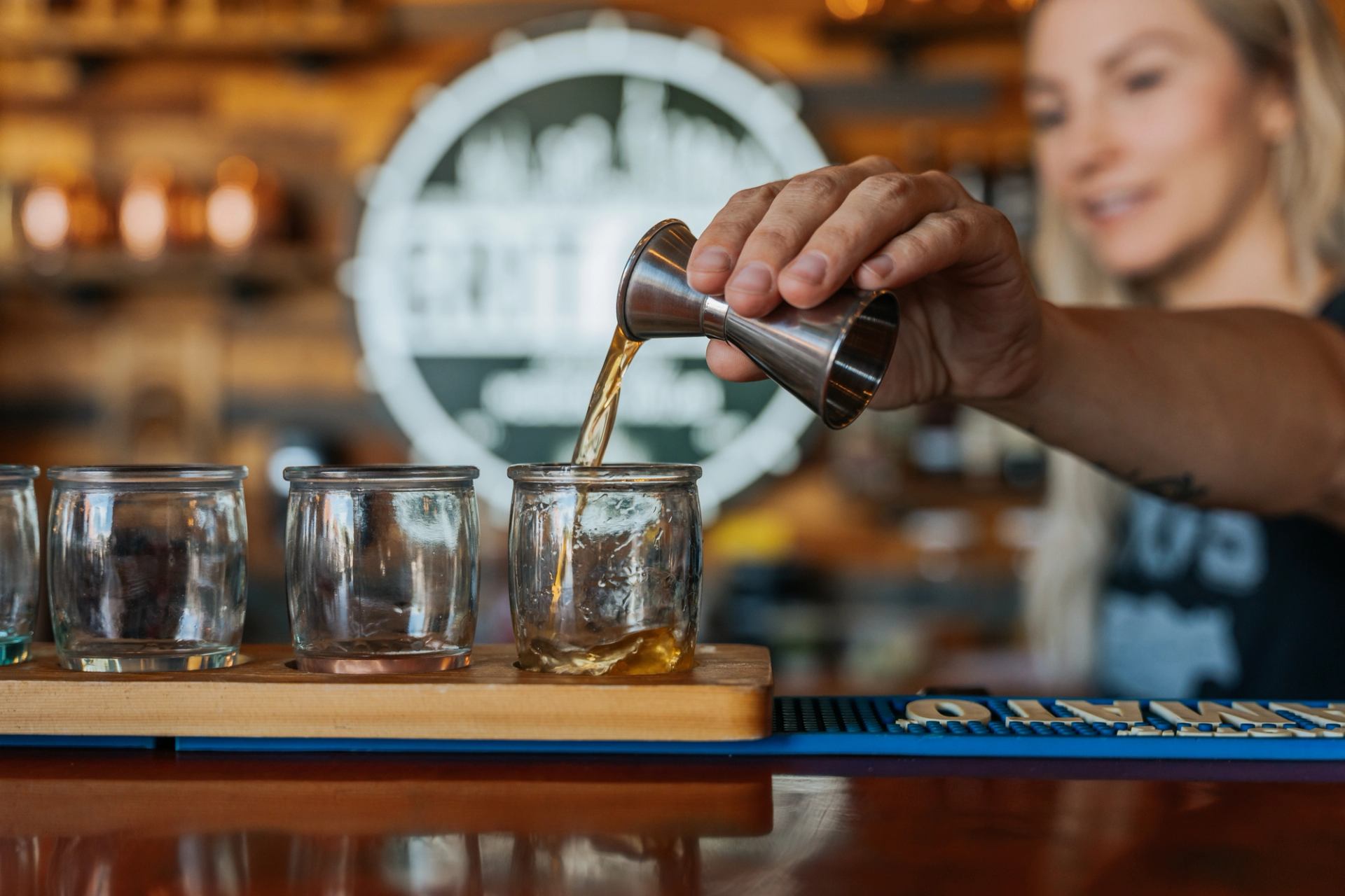 A drink being poured from a metal jigger into tasting glasses on a bar counter.
