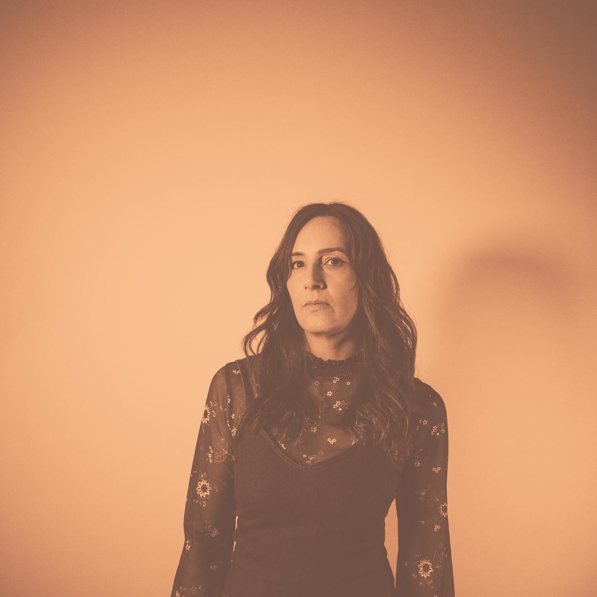 Person with long wavy hair stands against plain beige background in sepia tone.