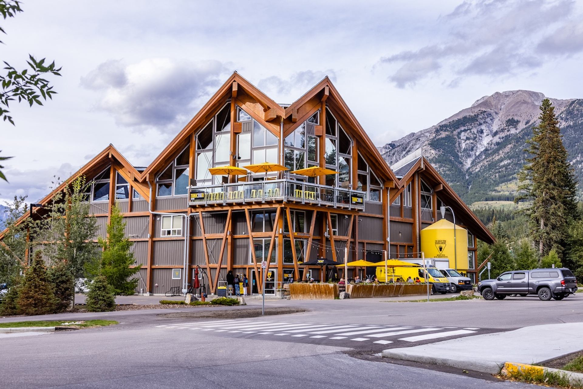 Grizzly Paw Brewery, a large lodge-style building with wood and glass, an outdoor patio with yellow umbrellas, and a yellow silo, against a mountain backdrop.