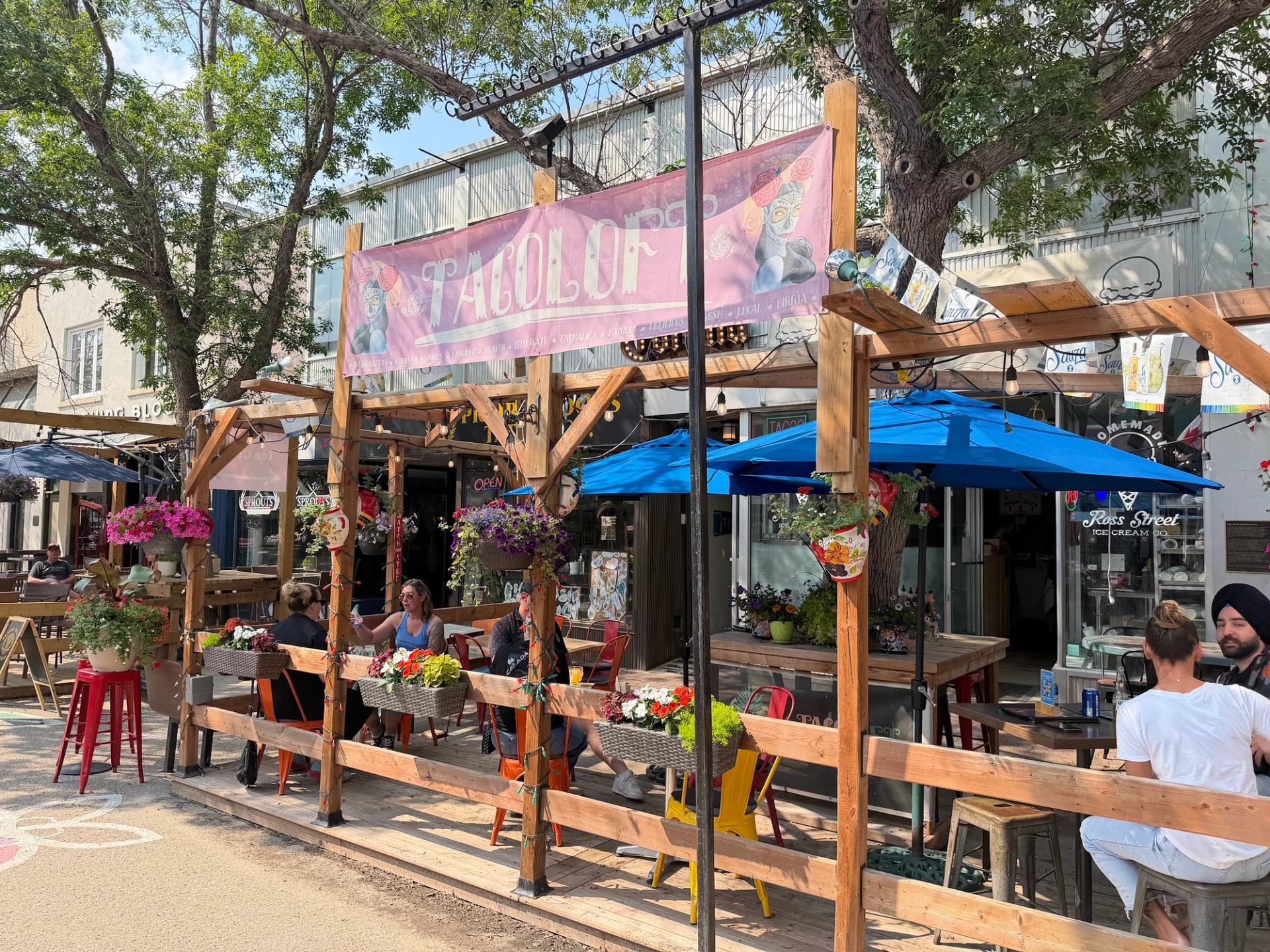 Street patio outside Tacolot restaurant with wooden pergola, blue umbrellas and hanging flowers.