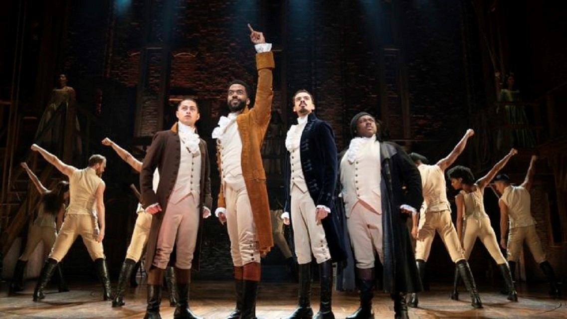 Theatre performers in period costumes bow on stage, one man with arms raised.