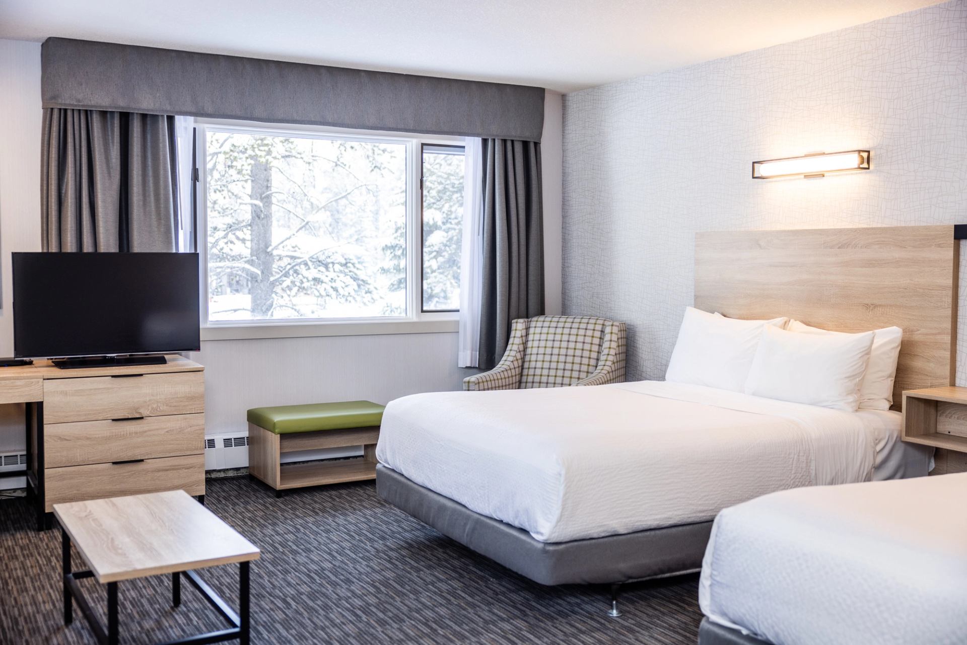 Bright hotel room with two beds, a TV, and a large window overlooking snowy trees.