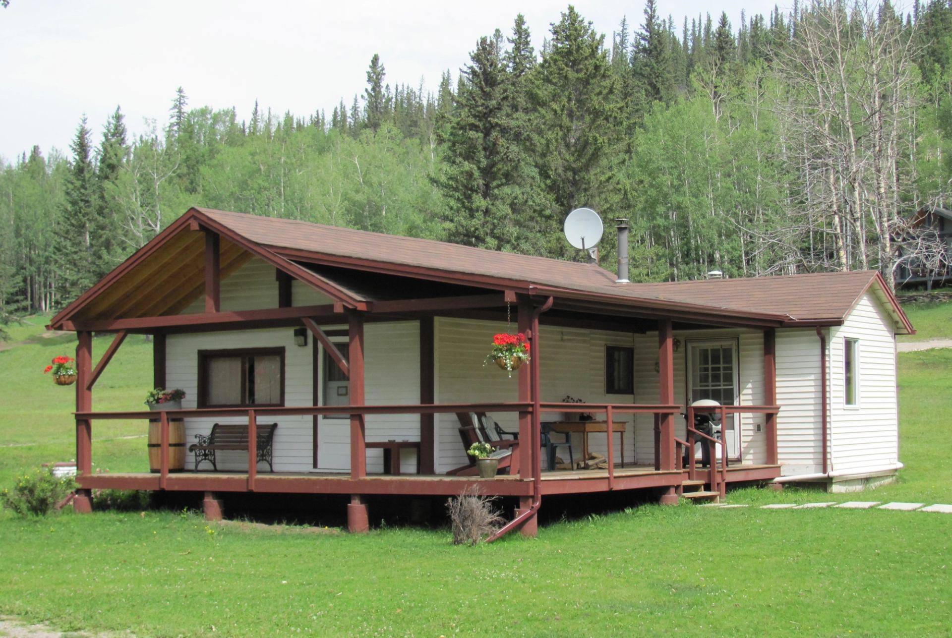 A white cabin with a brown roof and a wooden porch, surrounded by green grass and a forest.