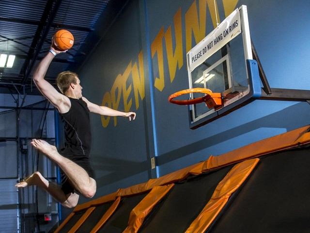 Man in black shirt dunking basketball at indoor trampoline park basketball hoop.