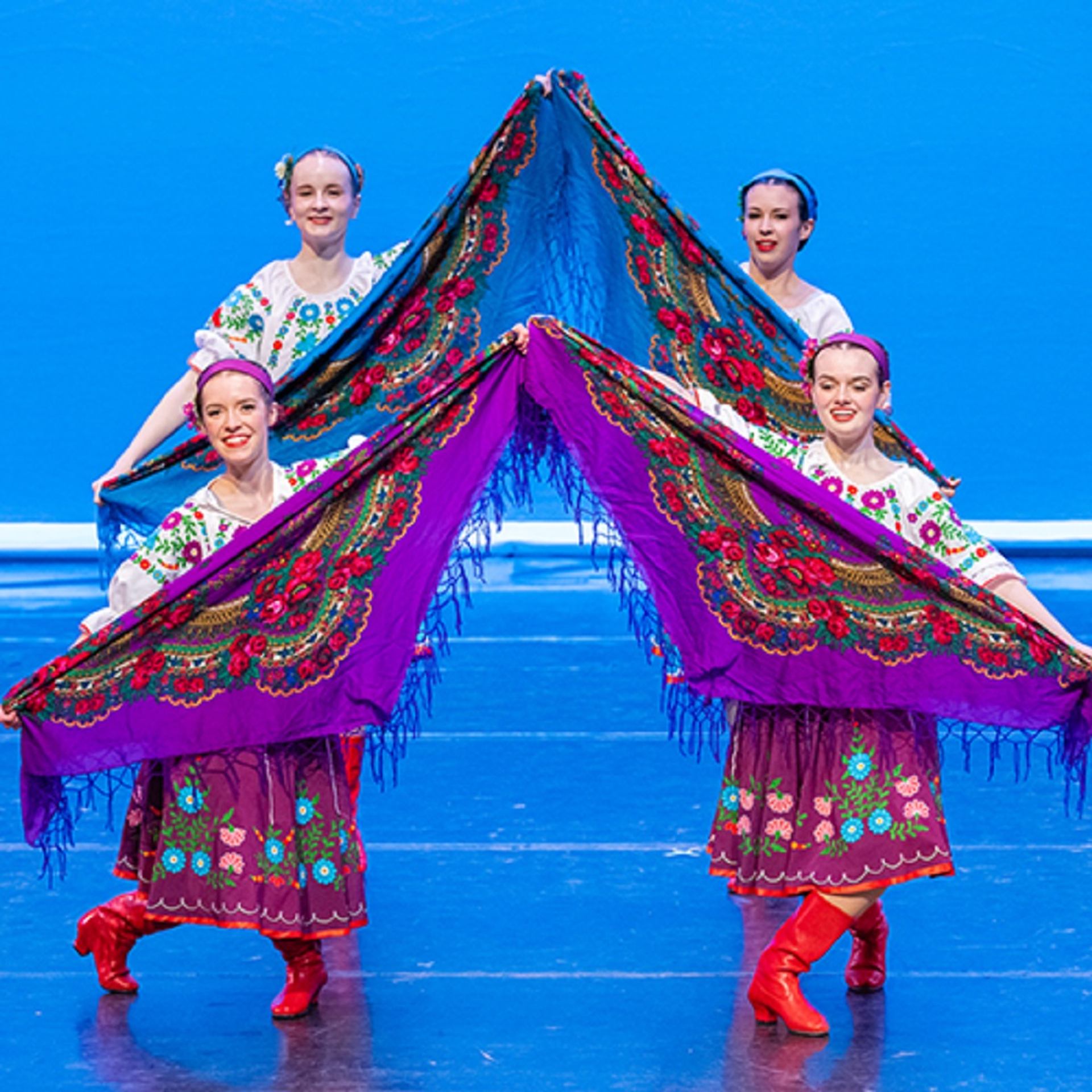 Dancers in vibrant Ukrainian folk costumes holding colorful patterned shawls on stage.