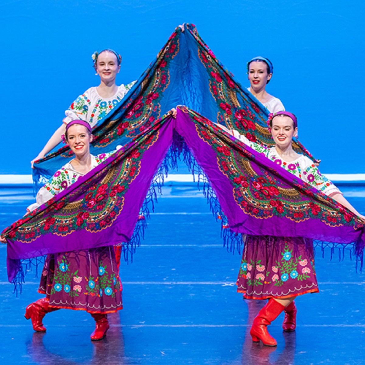 Dancers in vibrant Ukrainian folk costumes holding colorful patterned shawls on stage.
