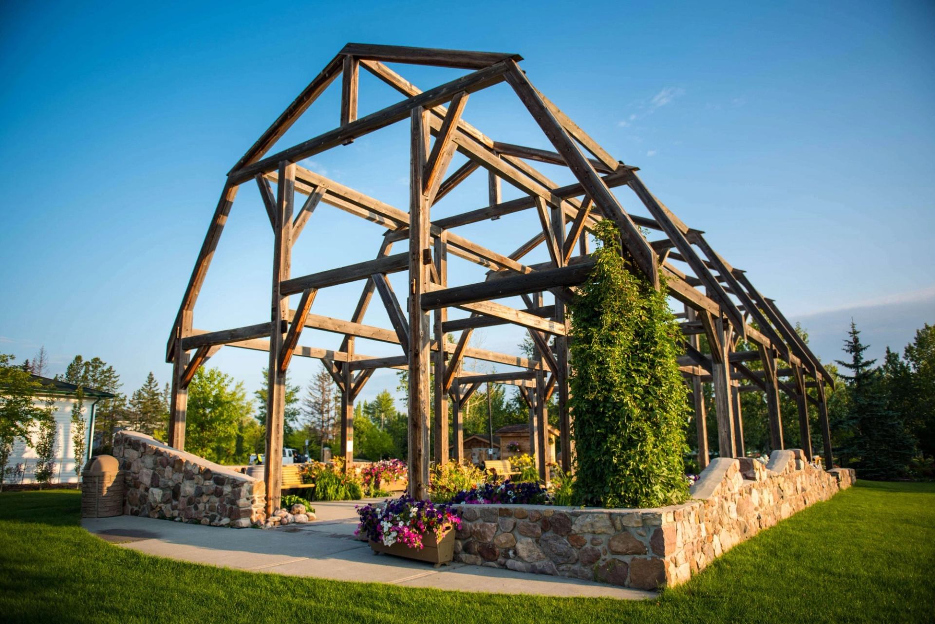 Open wooden pavilion frame with stone planters, green vines and colourful flowers.