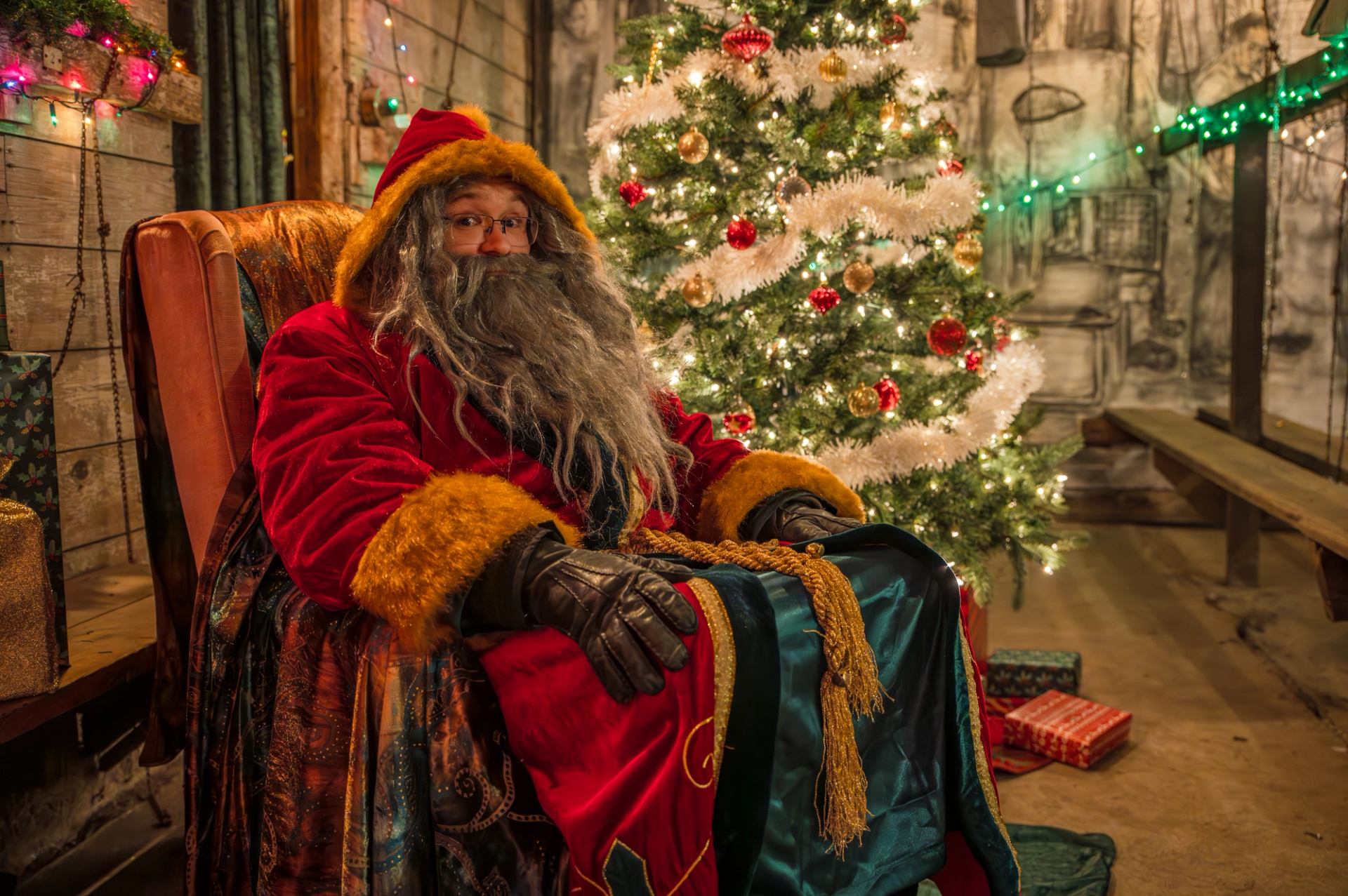A person dressed as Santa sits beside a decorated Christmas tree indoors.