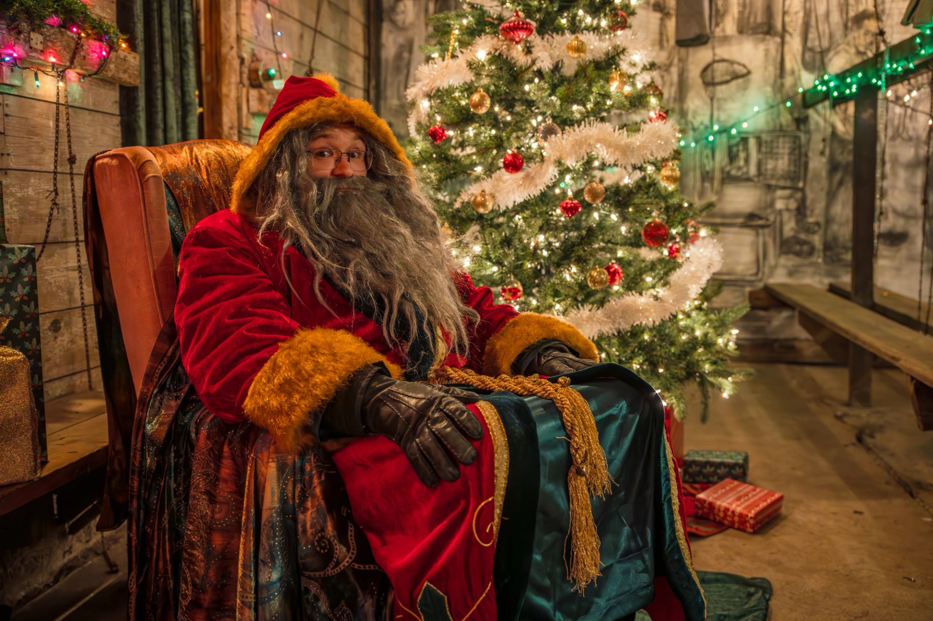 A person dressed as Santa sits beside a decorated Christmas tree indoors.