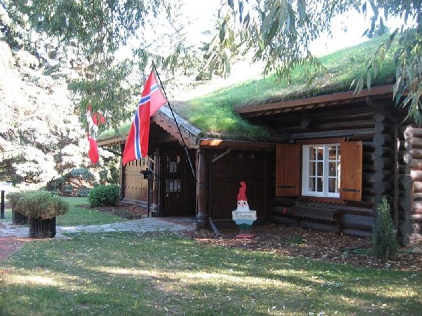 Norwegian log house with grass roof, flags, and gnome statue in a green park.