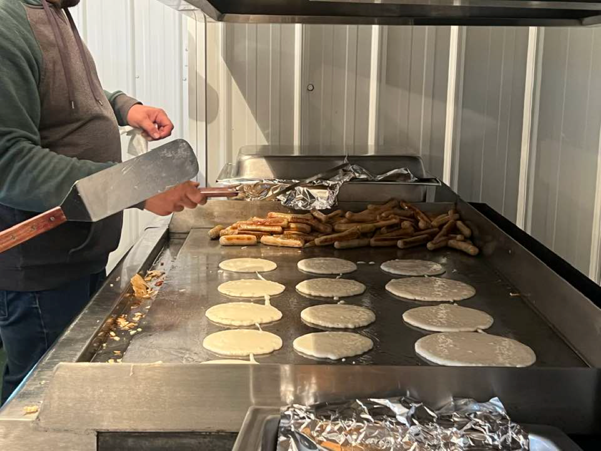 Person cooking pancakes and breakfast sausages on a large flat-top griddle indoors.