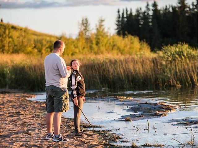 Two people standing on a sandy lakeshore at sunset, surrounded by reeds and calm water.